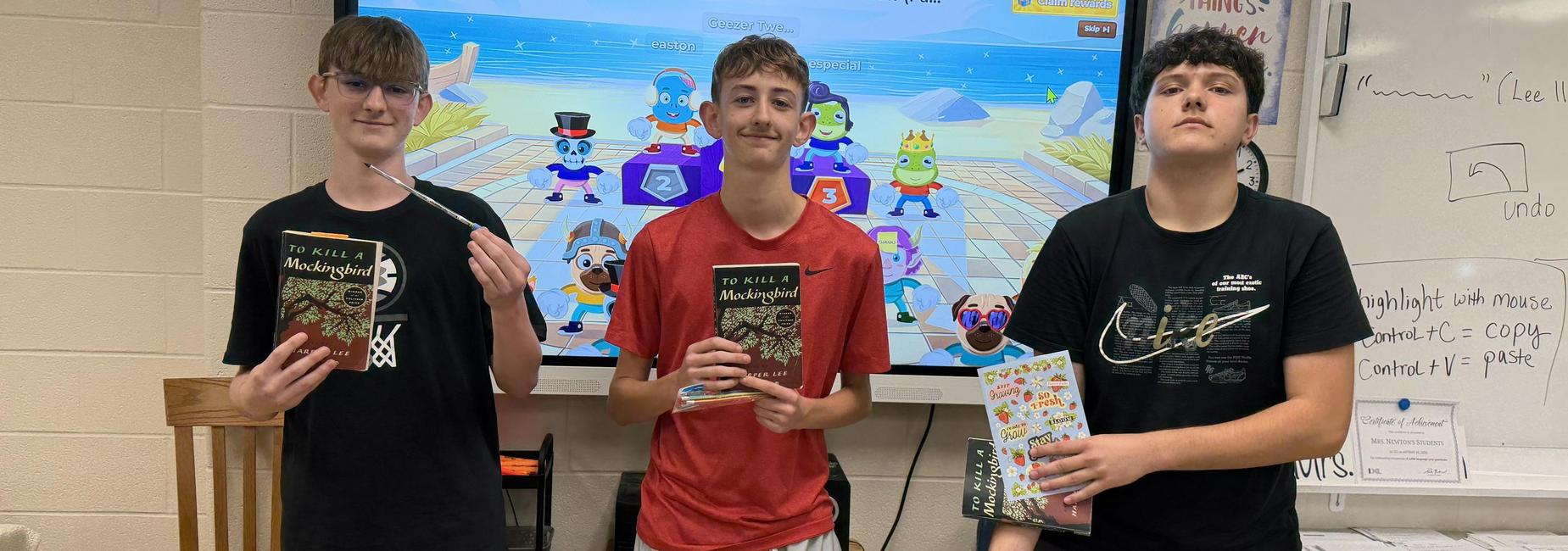 Three students holding books in a classroom with a colorful digital screen behind them.