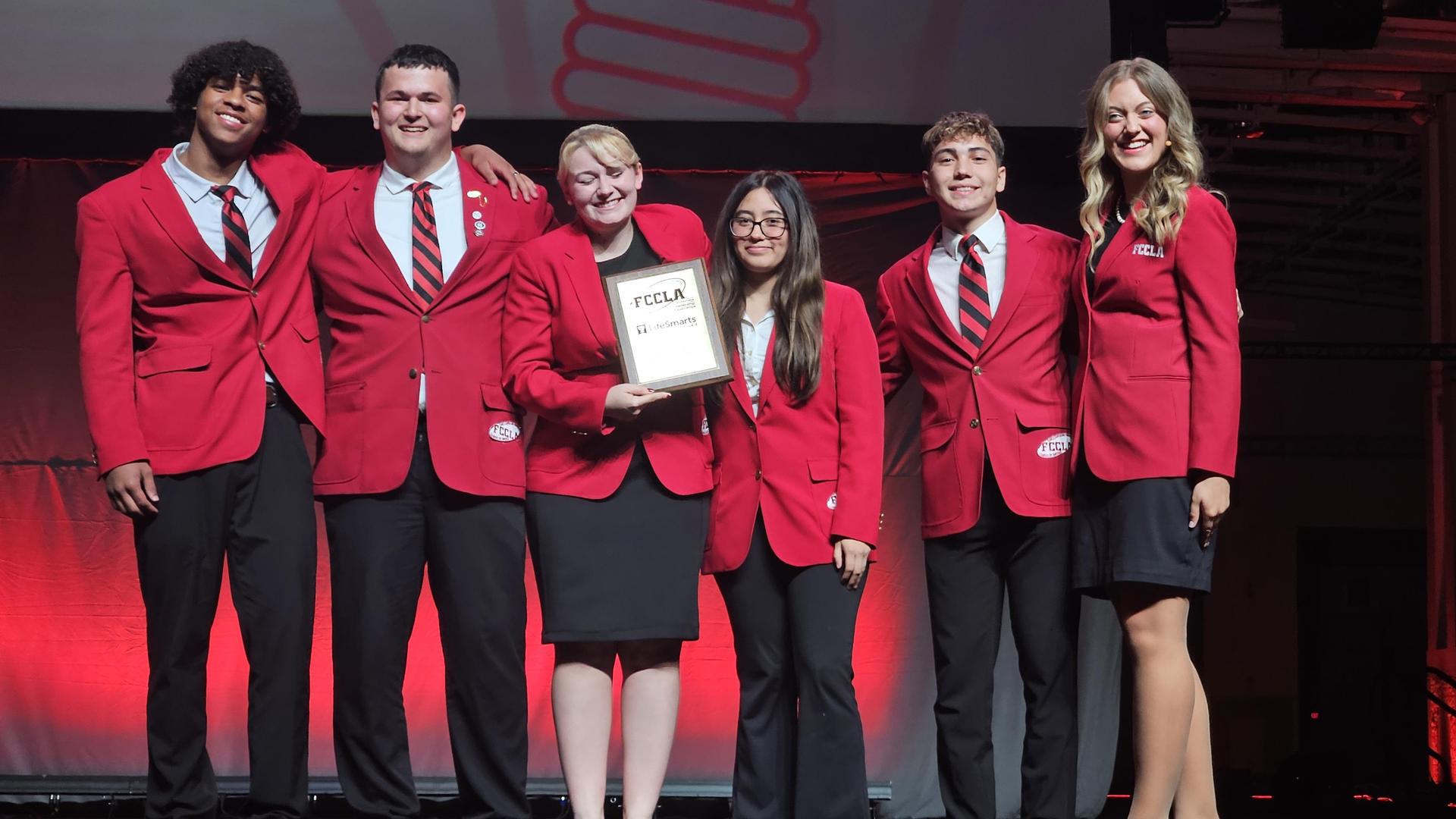 Group of six people in red blazers holding an award on stage.