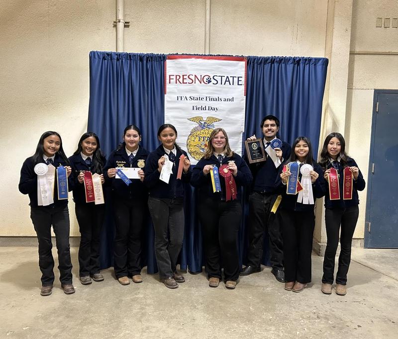 Eight FFA students stand in front of a banner reading “Fresno State FFA State Finals and Field Day,” proudly displaying an array of ribbons in various colors. One student in the center holds a plaque, celebrating the group’s strong performance.