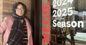 A young woman wearing glasses, a striped top, and a pink coat stands outside Carnegie Hall next to a window decal that reads “2024–2025 Season.” Snow is visible on the ground inside the reflection, marking the winter setting of the moment.