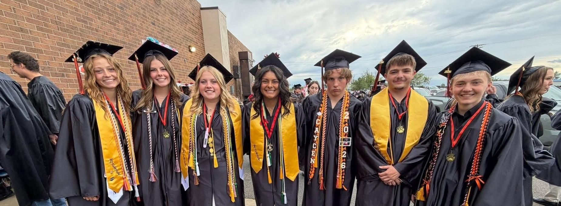 Group of seven graduates in caps and gowns showcasing their honors and smiles.