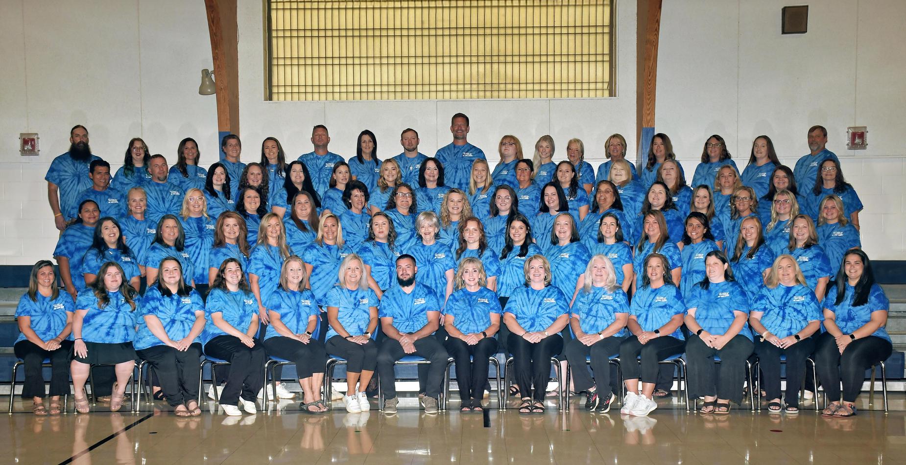 Group photo of a large team wearing blue tie-dye shirts, smiling and seated in rows.