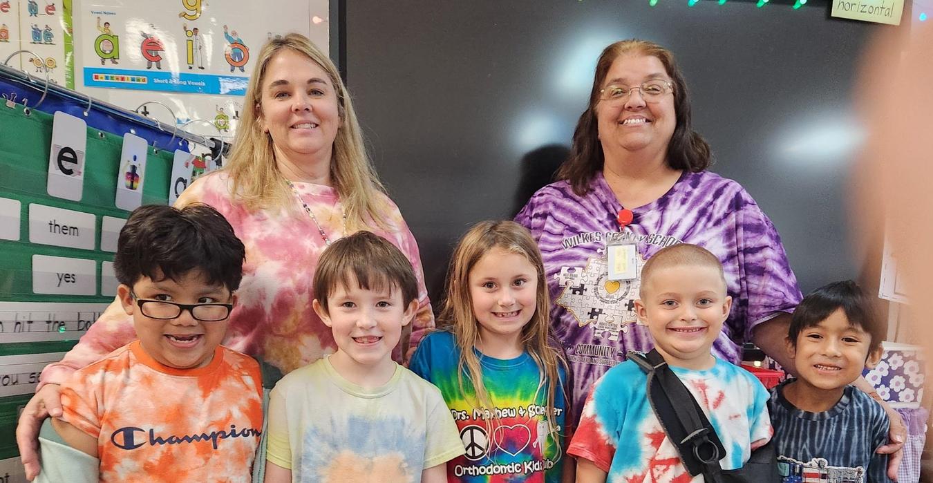 A group of five children with colorful tie-dye shirts smiling together in a classroom setting.