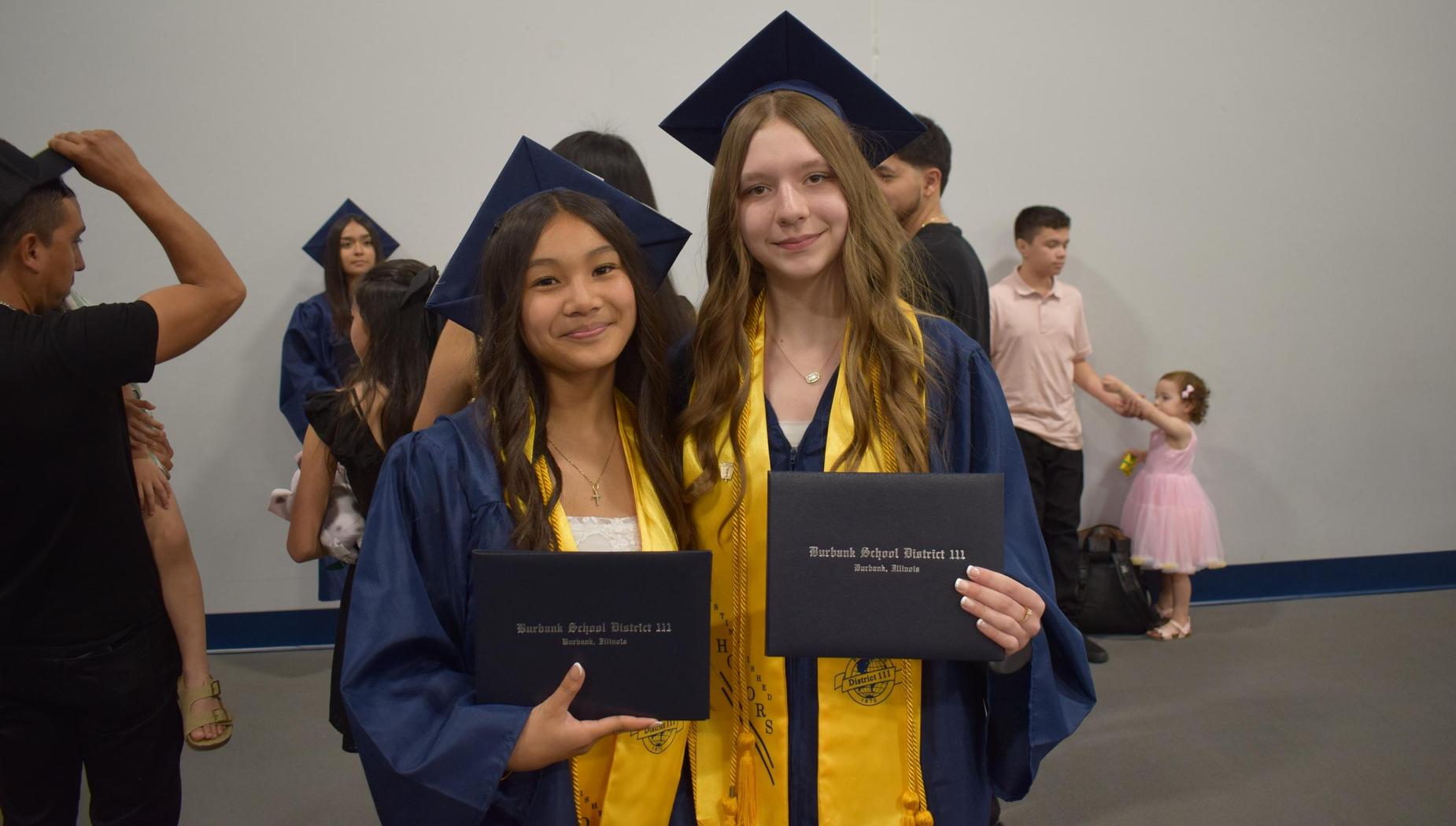 Two graduates pose with diplomas, surrounded by other graduates.