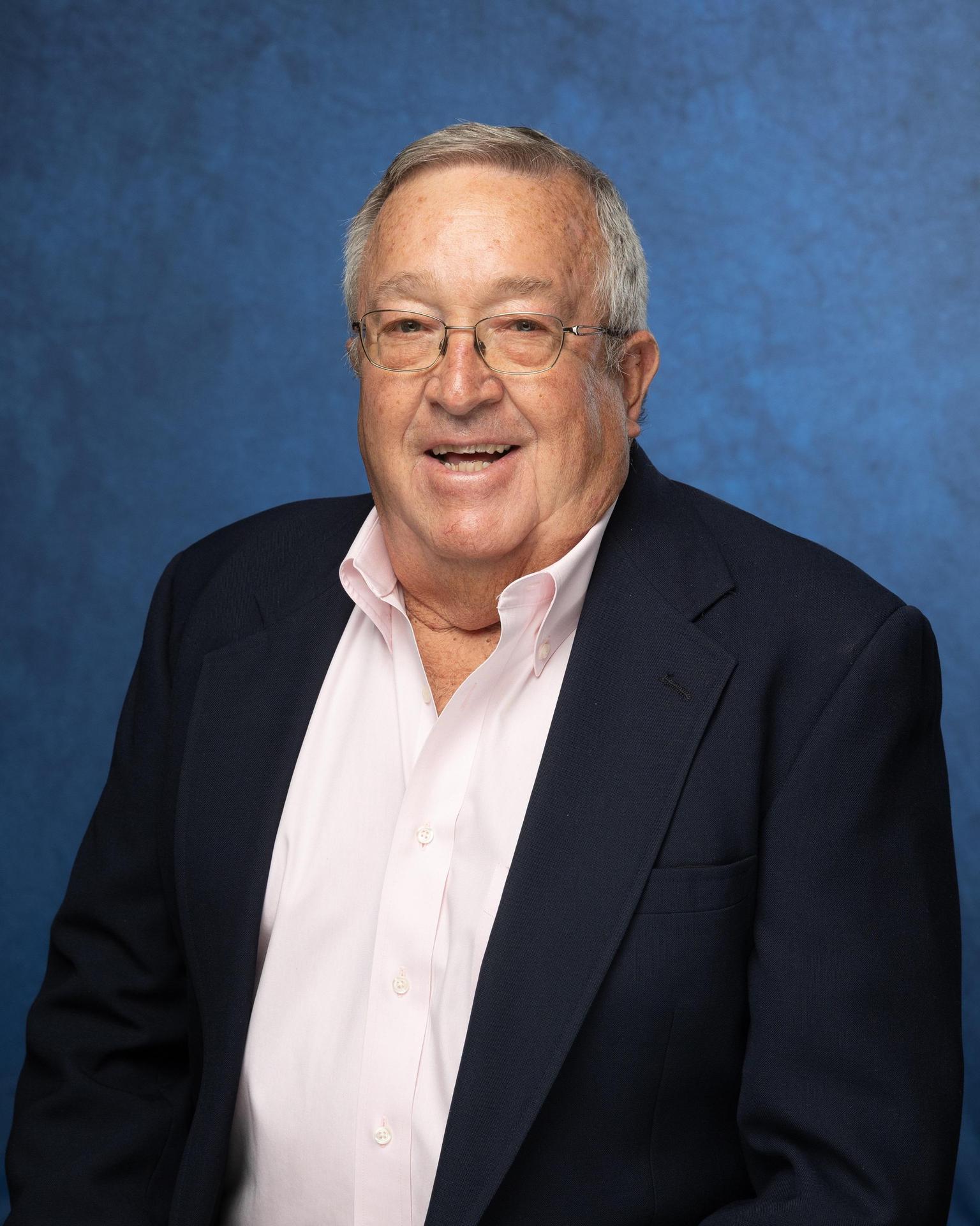 Smiling older man in glasses wearing a dark suit and pink shirt against a blue background.