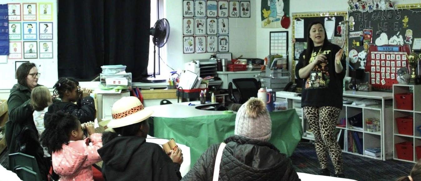 A teacher wearing leopard print pants presents to a classroom of seated students and parents.