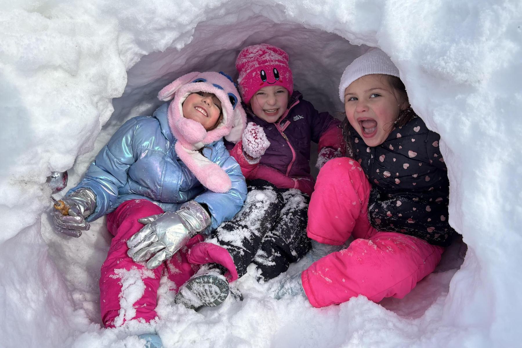 Three children smiling and playing inside a snowy fort.