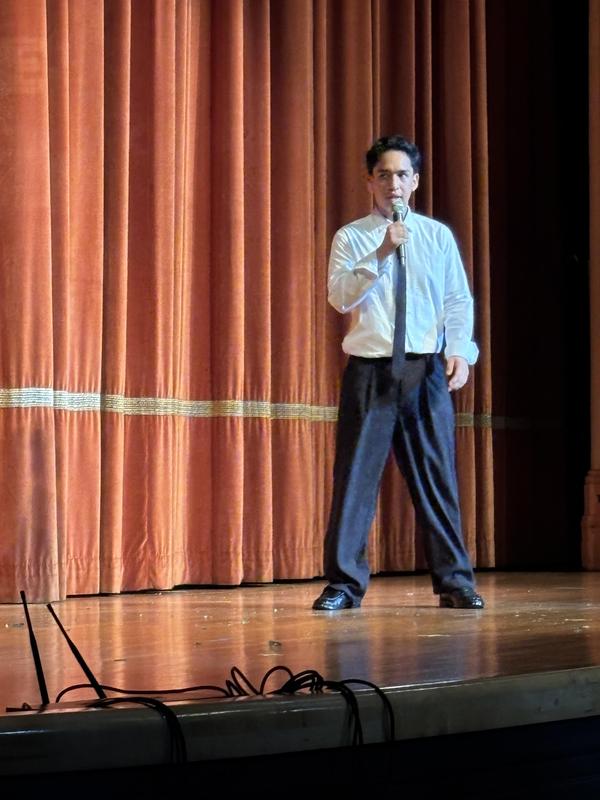 Man in a white shirt and tie holding a microphone on stage before orange curtains.