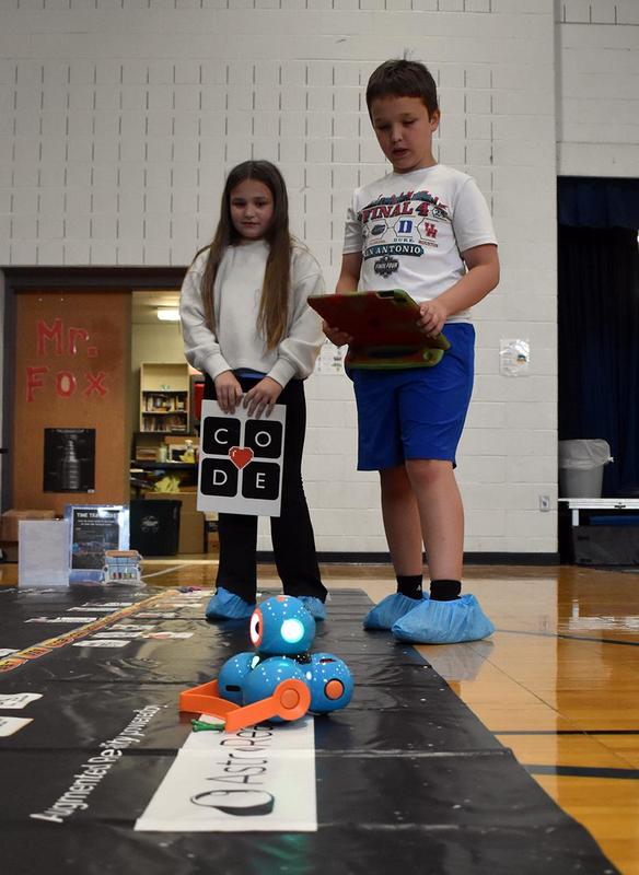A girl holds a 'CODE' sign while a boy operates a robot with a tablet.