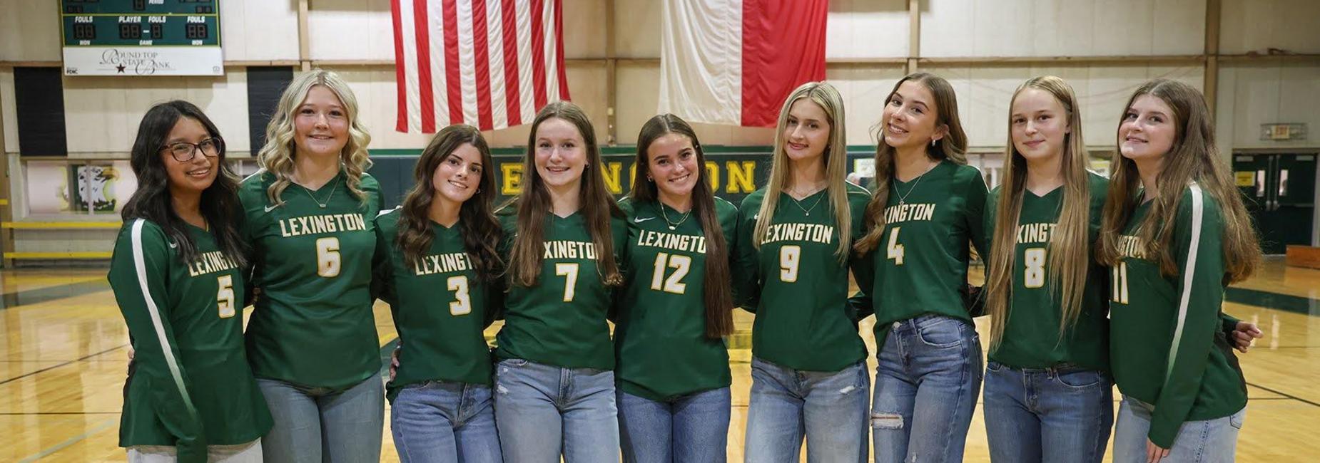 Eight individuals in green jerseys and jeans, posing in a gym with a sports court.