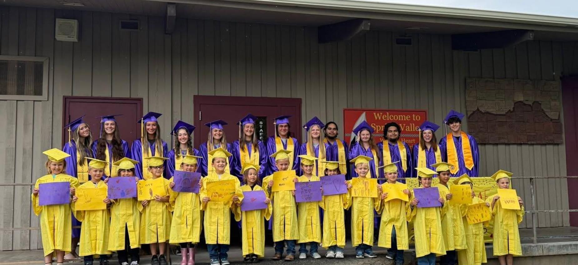 Spring Valley graduates in yellow and purple gowns holding signs outside a building.