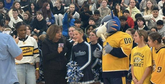 cheerleader talking into a microphone being filmed for a tv show standing with a mascot dressed in nashville predators hockey gear with students sitting in the bleachers behind her