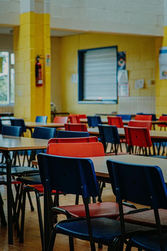 School Cafeteria with empty tables and red and blue chairs arranged in rows.