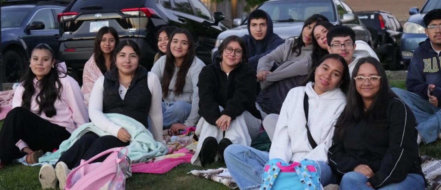 A group of teenagers sitting on the grass with blankets and backpacks, smiling.