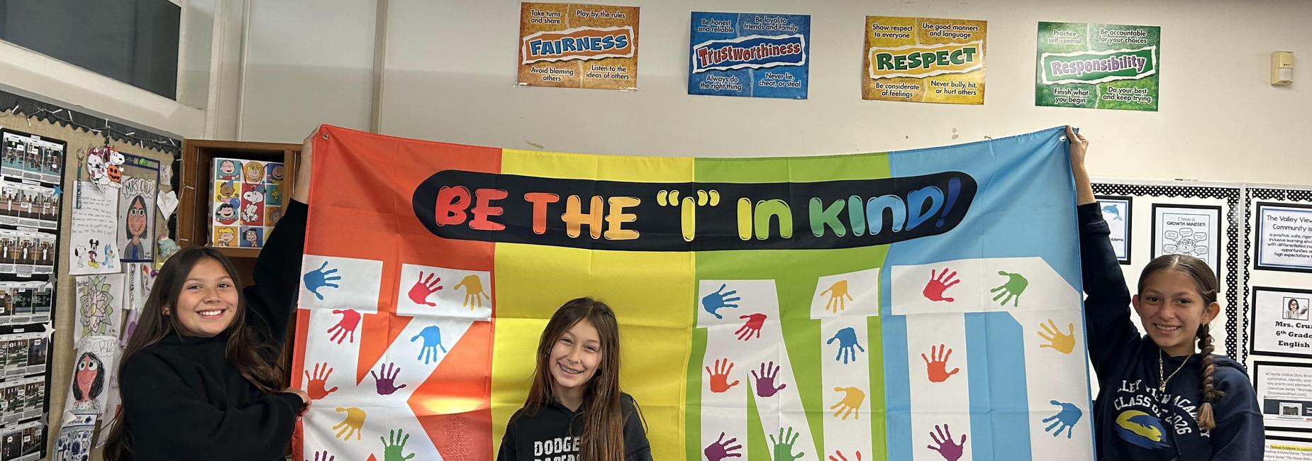 Three girls holding a large colorful banner promoting kindness.