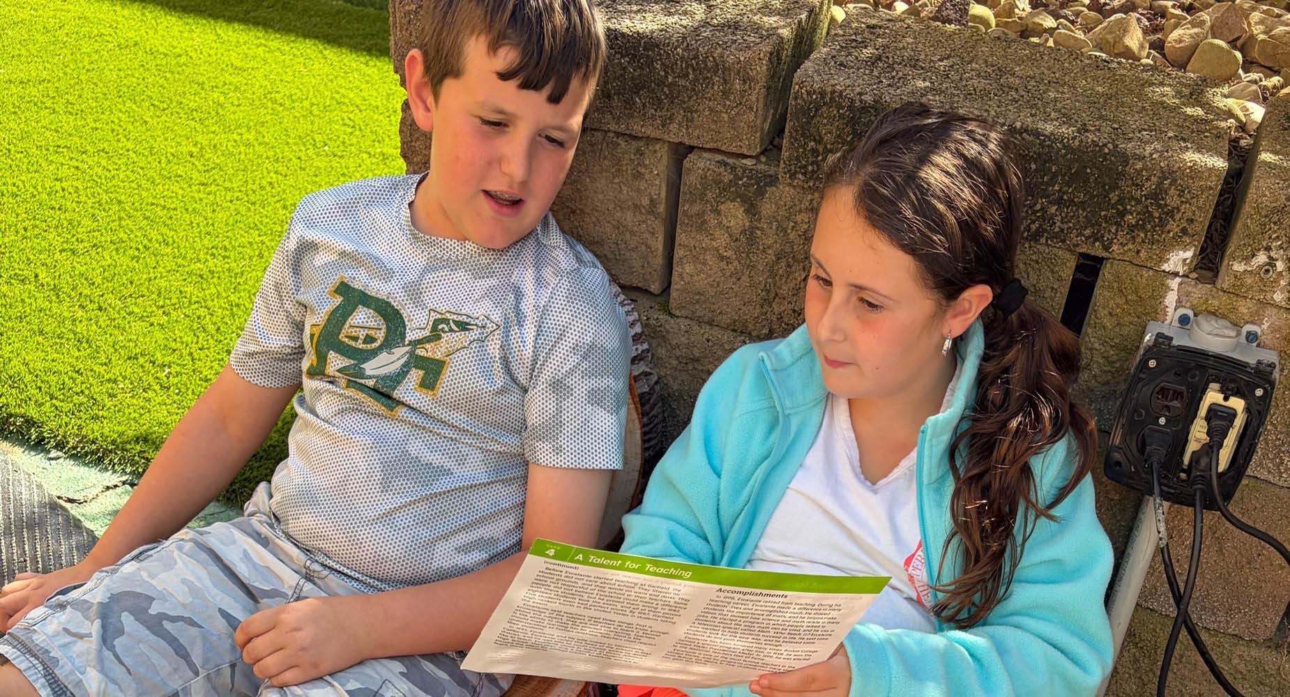 Two children sitting outside, reading a document together.