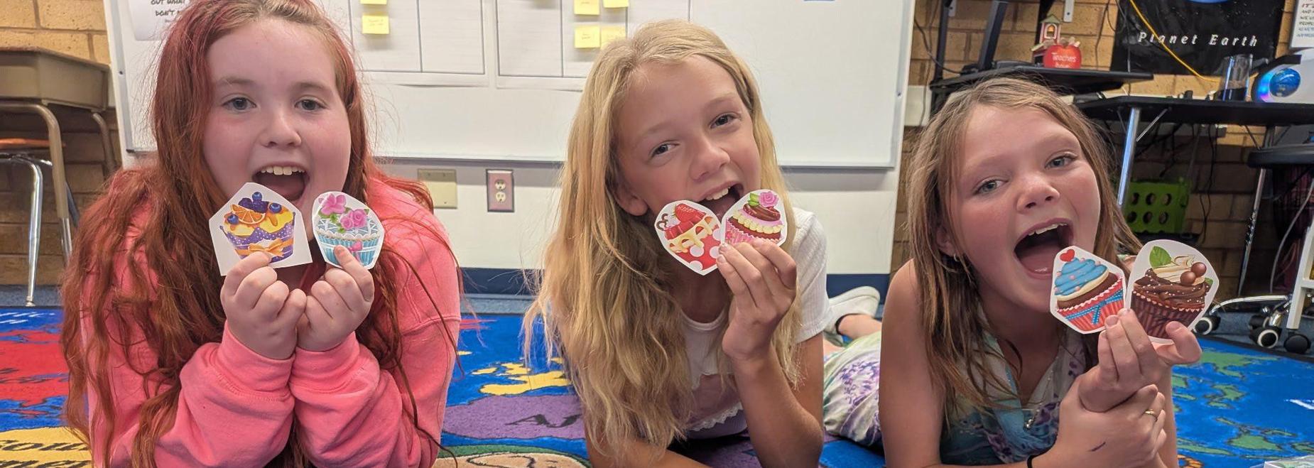 Three children hold decorated cupcake cutouts while sitting on a colorful classroom rug.