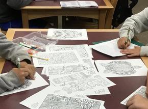 Classwork pages scattered across a table with hands from multiple students reaching in and writing collaboratively