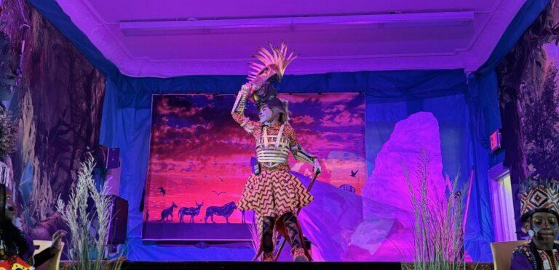 A performer in elaborate costume dances on stage with colorful background scenery.