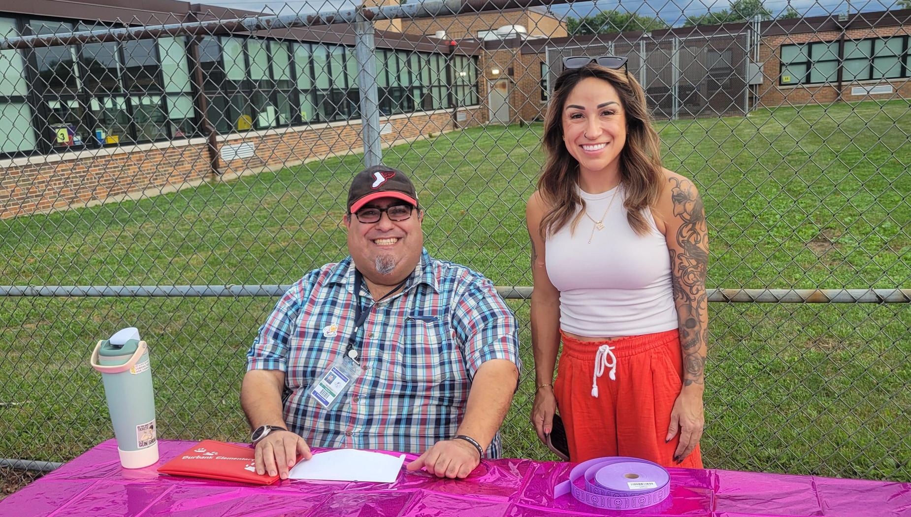 Two smiling people at a table with a pink tablecloth in an outdoor setting.