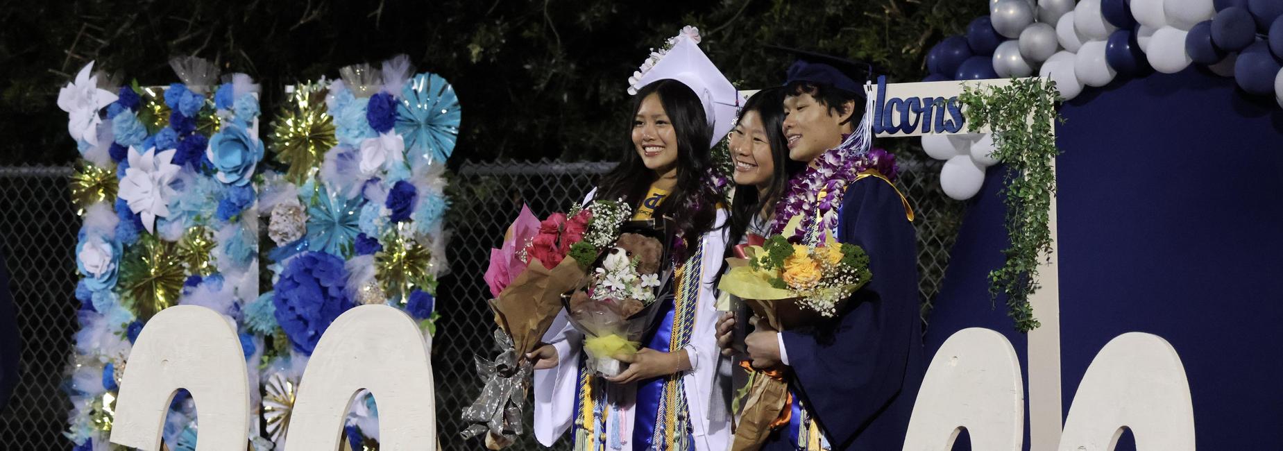 Two graduates pose together with bouquets beside a large '2026' sign.