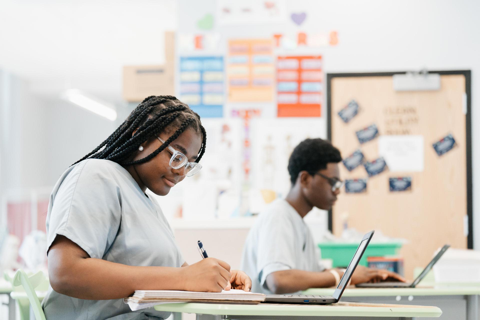 Students work at desks, one writing in a notebook and another using a laptop in a classroom.