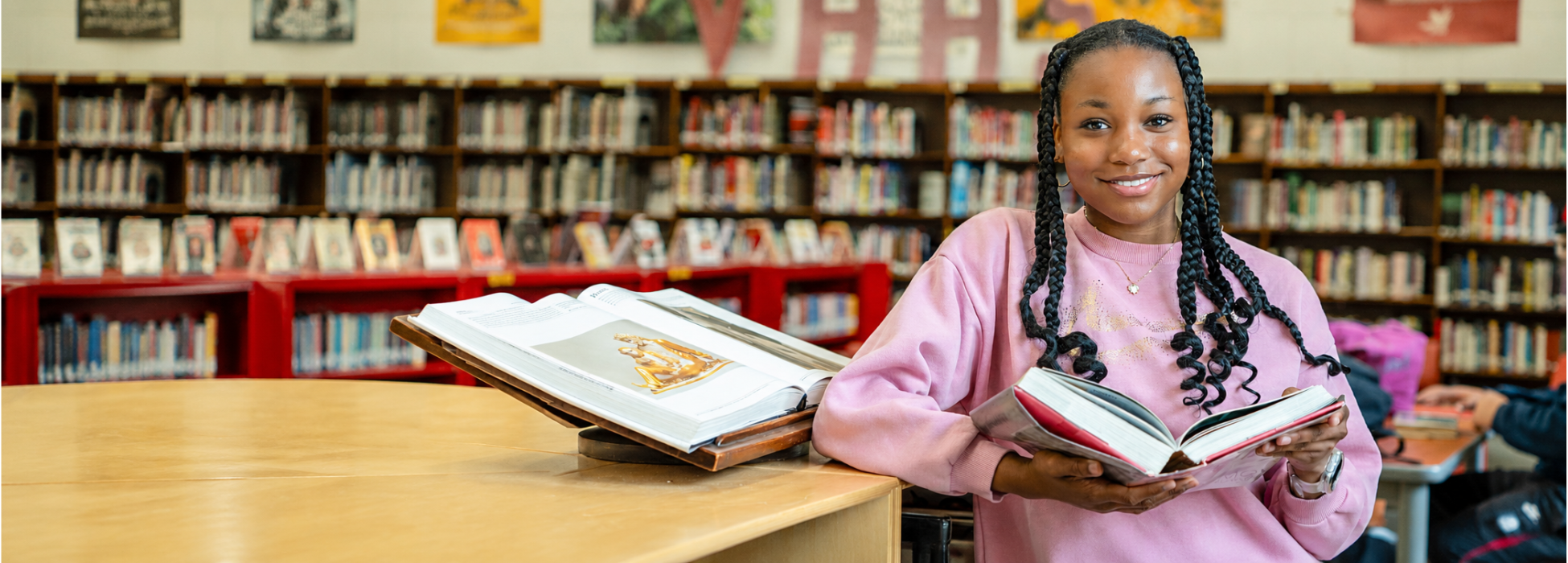 A smiling girl holds a book in a library with many shelves in the background.
