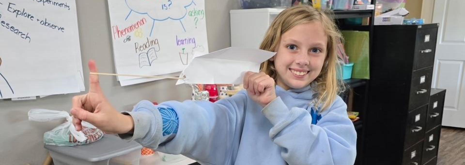 Child holding a craft project with a big smile in a classroom.