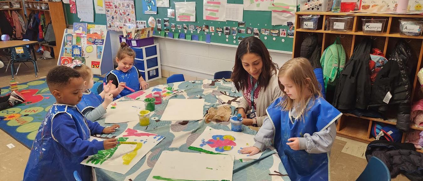 Children are at a table wearing blue aprons painting pictures while a teacher is looking on.