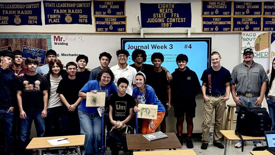 Group of students and teacher posing in a classroom with artwork and a presentation behind.