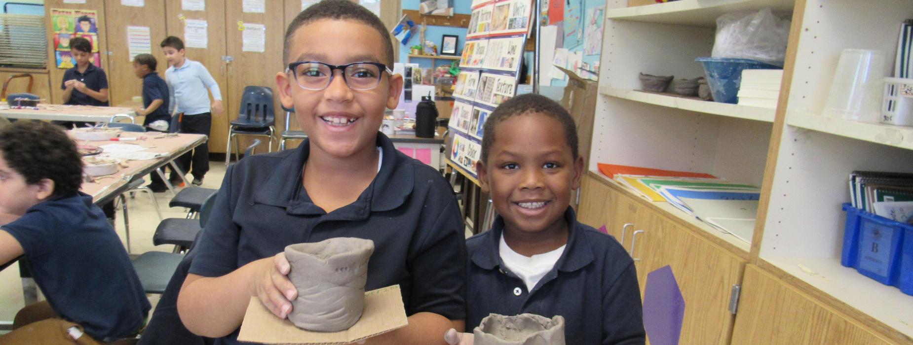 Two smiling boys holding clay pots in a classroom.