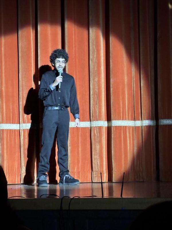 A performer with curly hair singing into a microphone on stage against a curtain backdrop.