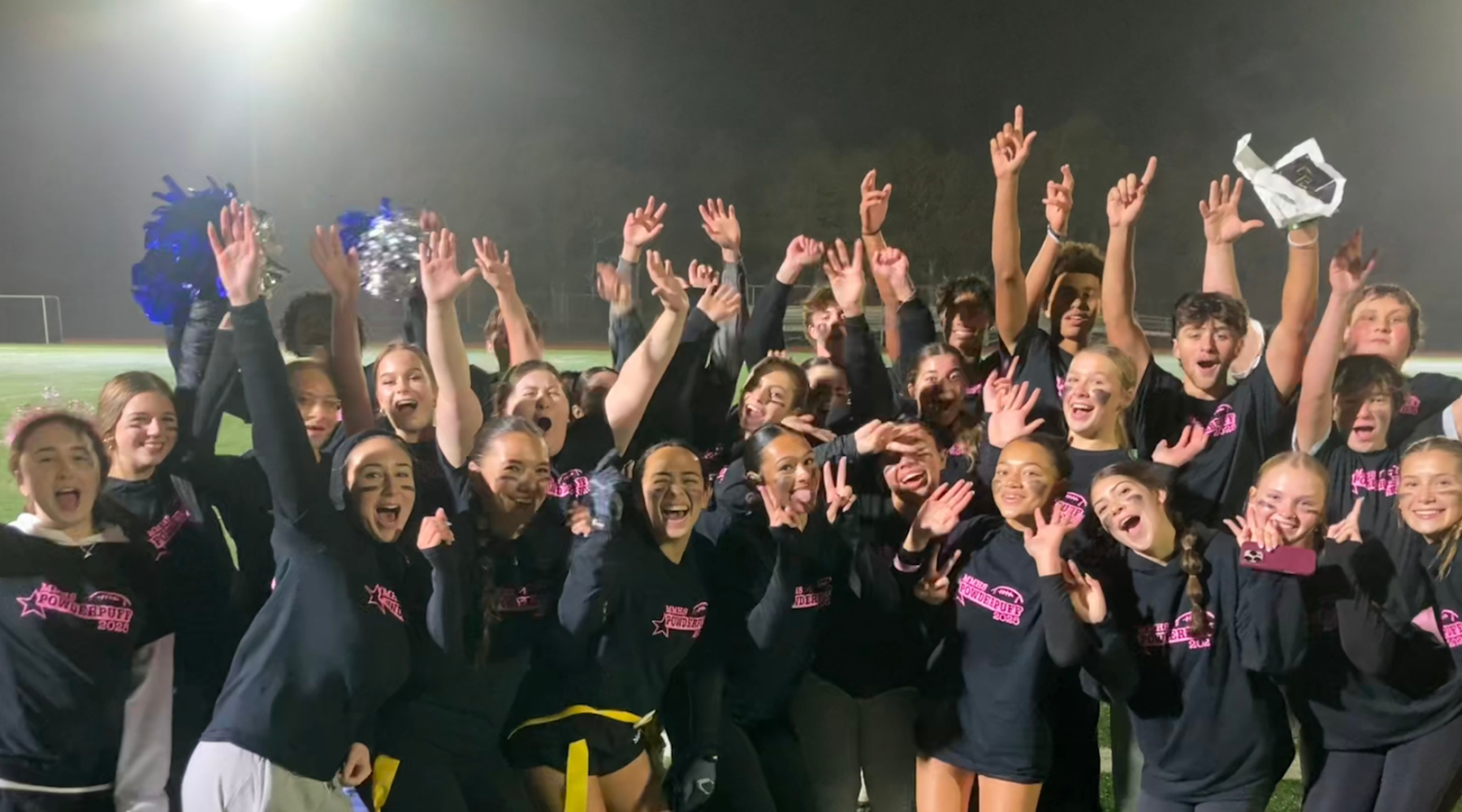 Cheerleaders celebrate in a group, raising their arms in excitement at night.
