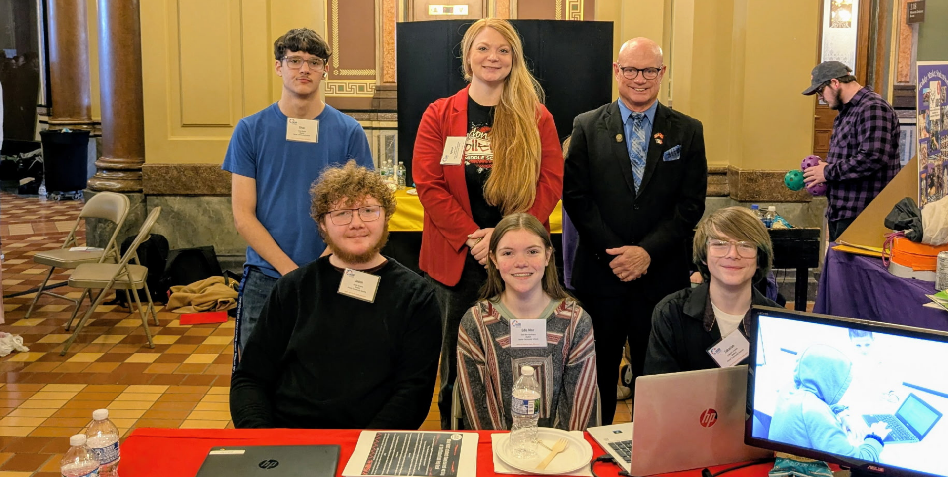 Five people stand and sit behind a table at an event, featuring laptops and various displays.