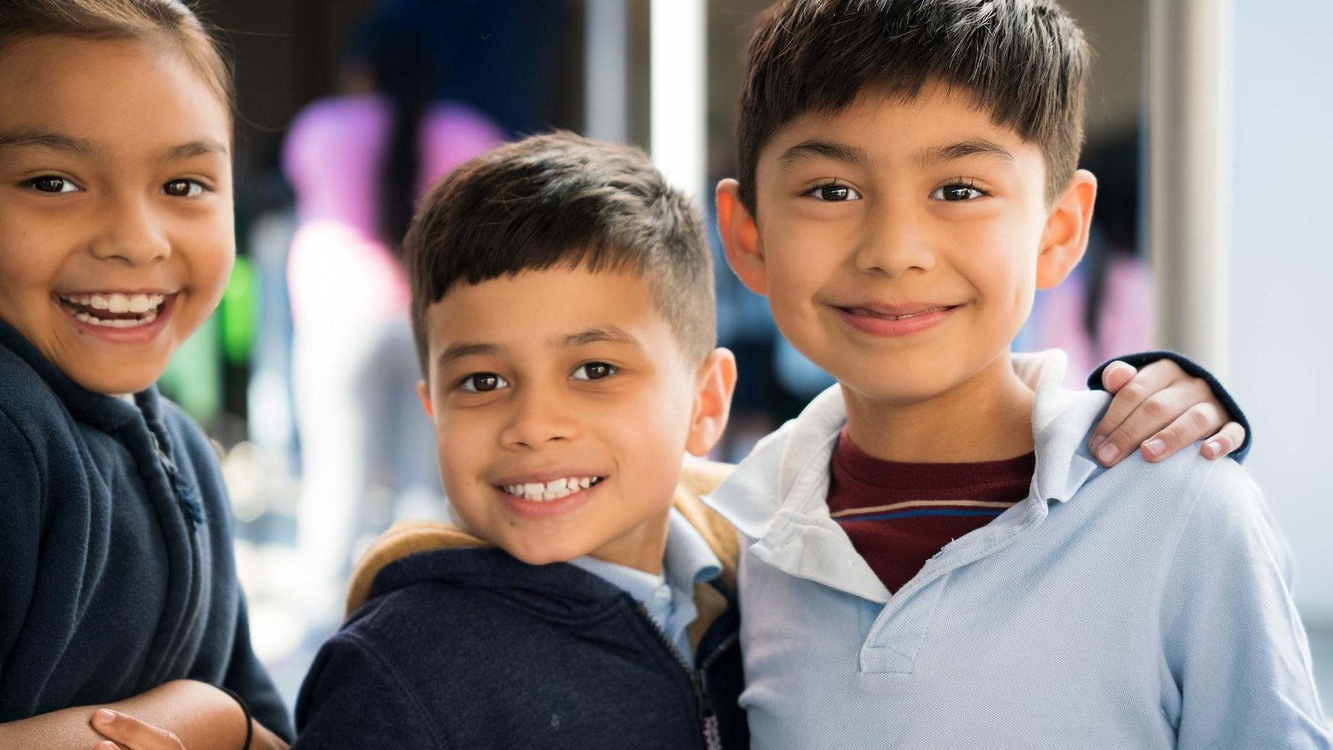 Three happy children pose together, grinning in a bright setting.