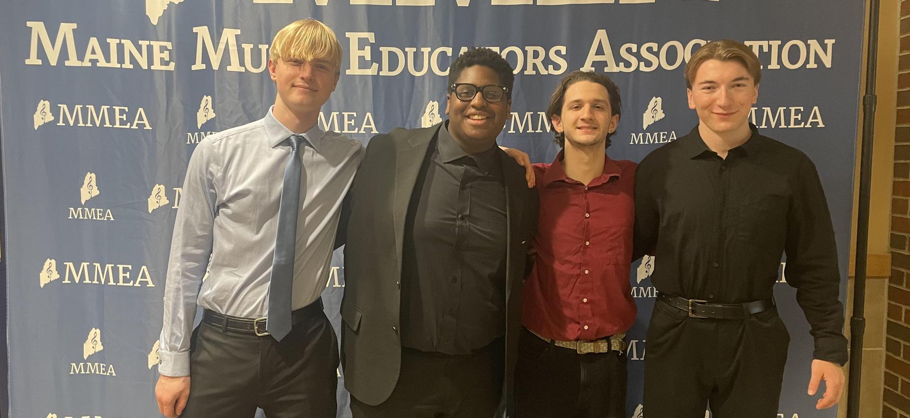 Four young men dressed in formal attire stand together in front of a blue banner.