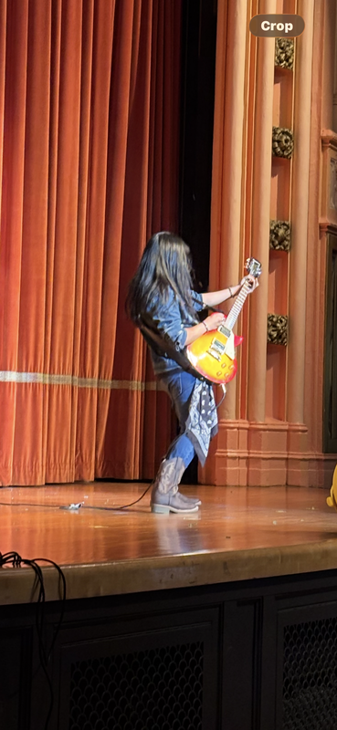 Person playing an electric guitar on a stage with orange curtains.