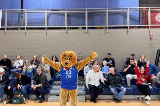 A lion mascot poses with arms raised in a gym, while an audience sits in the background.