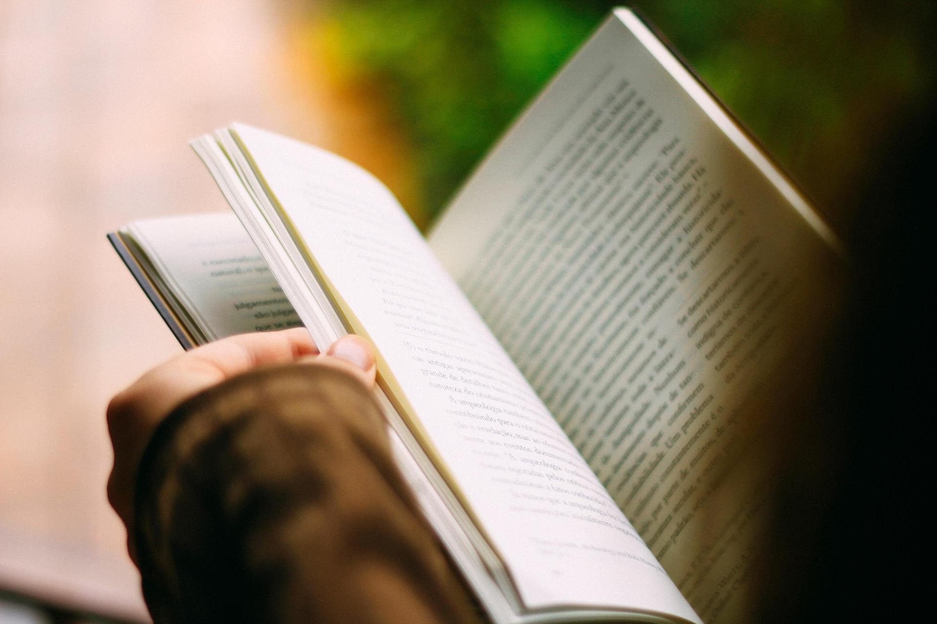 A person holding an open book with a blurred green background.