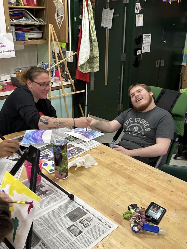 An instructor assisting a student with their painting at a communal art table.