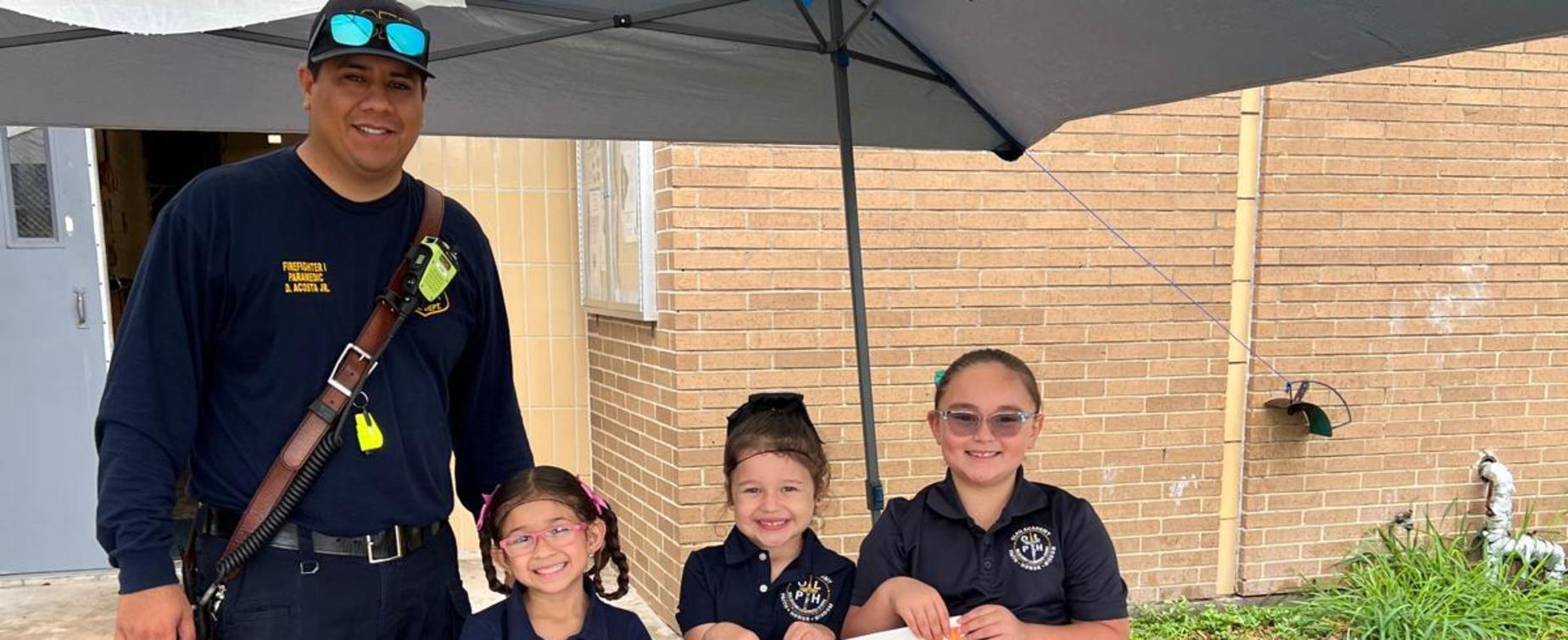 A police officer stands with three girls holding a sign for a bake sale outside a school.