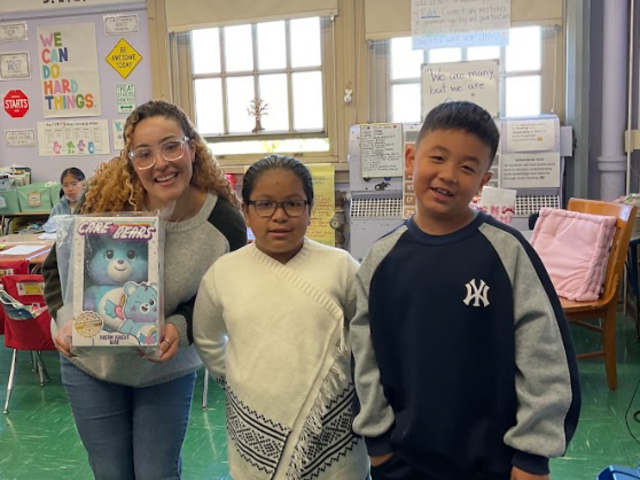 Three children and an adult posing together in a classroom, smiling and holding a toy box.