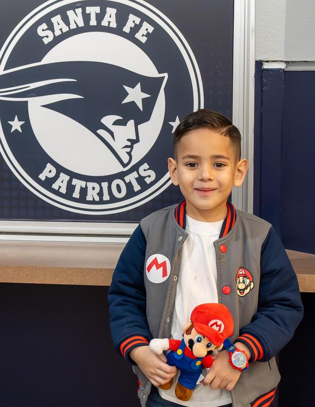 A young student stands in front of a Santa Fe Patriots logo, smiling and holding a Super Mario plush toy. He wears a Mario-themed jacket, watch, and T-shirt, proudly showing off his matching outfit.
