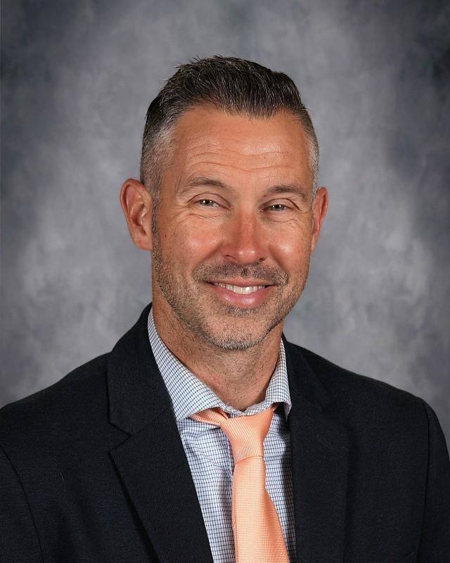 Smiling man in a gray suit with a plaid tie against a gray backdrop.