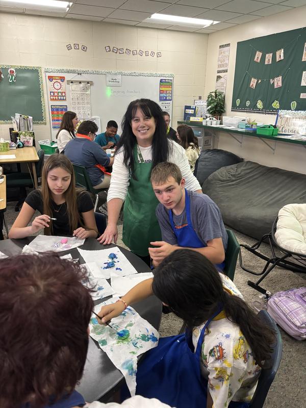 A teacher interacts with students painting at tables in an art classroom.