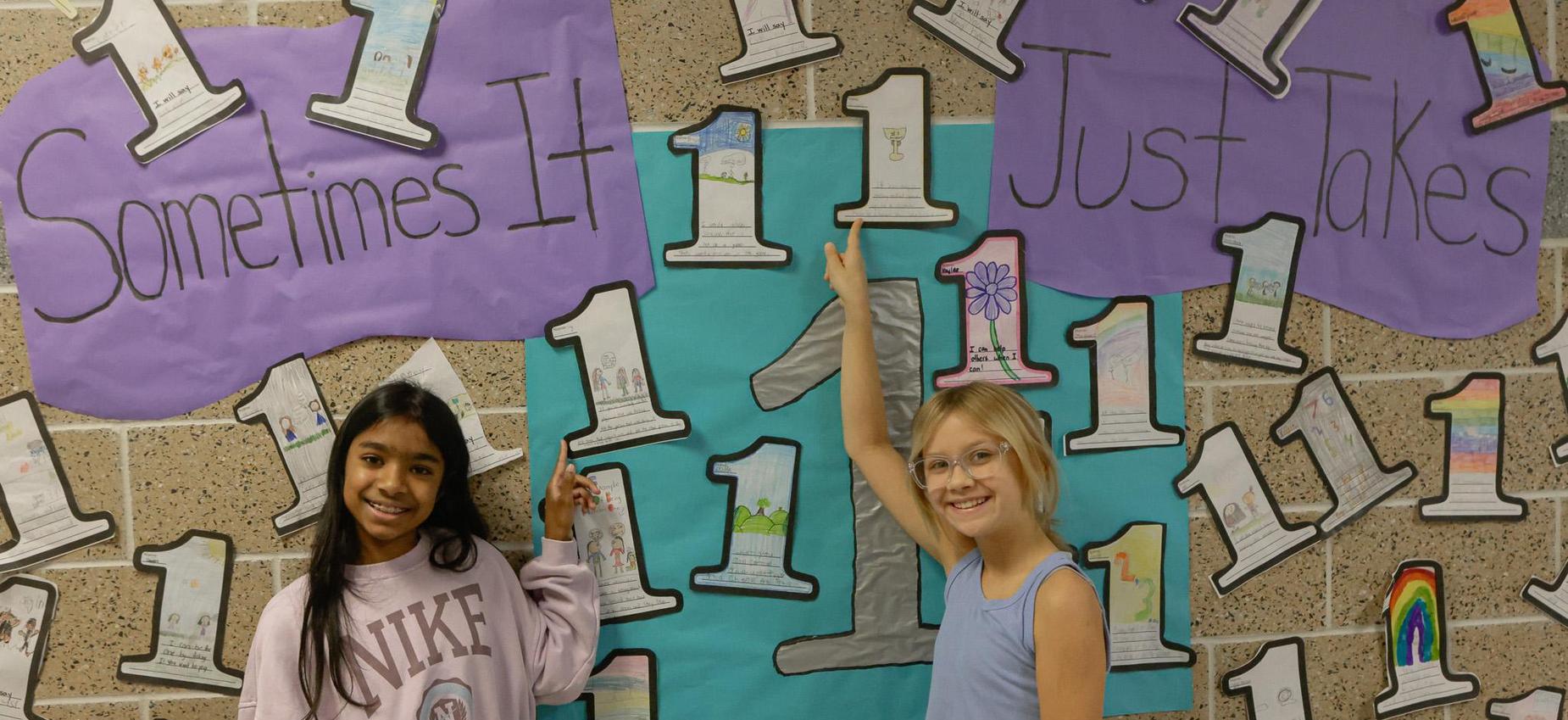 Two girls point at a mural with the text 'Sometimes It Just Takes 1' and colorful drawings.