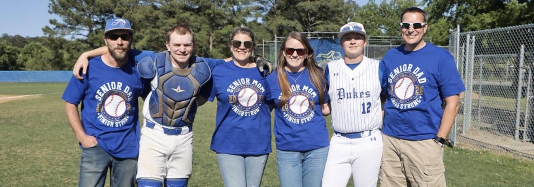 A group of five people wearing blue shirts, posing on a baseball field.