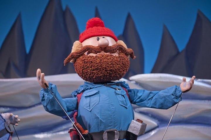 A bearded puppet wearing a red beanie and blue jacket stands on a snowy terrain.