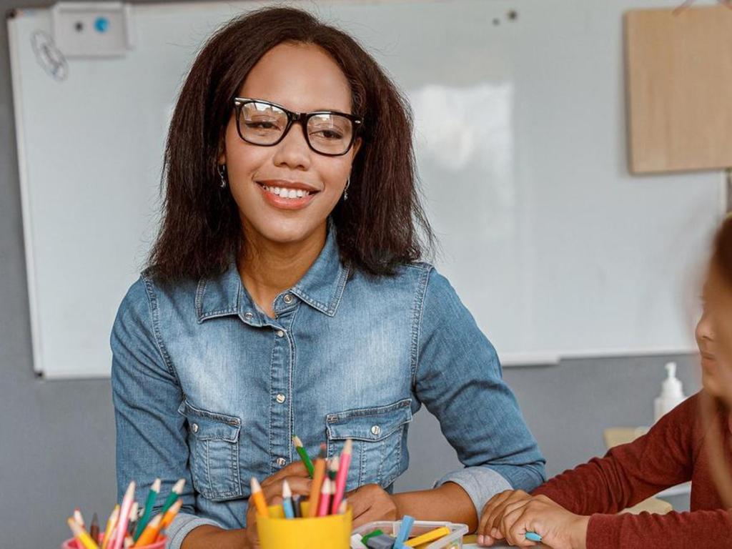 A smiling woman with glasses sitting at a table with colorful art supplies.