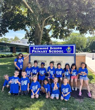 Group of children in blue shirts posing in front of a school sign.